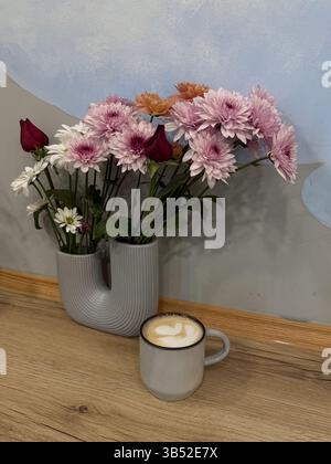 Cette photo capture une tasse de cappuccino en céramique aux tons neutres avec de la mousse de lait placée sur un plancher en bois à côté d'un vase en céramique de forme unique tenant un Banque D'Images