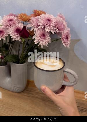 Cette photo montre un gros plan d'une main tenant une tasse en céramique remplie de cappuccino surmontée de mousse de lait en forme de cœur. Derrière la tasse se trouve un décoratif Banque D'Images