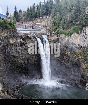 Vue panoramique sur les chutes de Snoqualmie cascadant sur une falaise rocheuse avec une centrale hydroélectrique et des collines boisées en arrière-plan. Banque D'Images