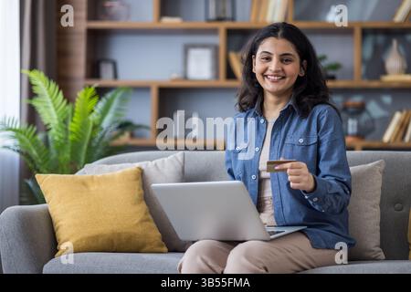 Portrait d'une jeune femme indienne souriante dans une chemise assise sur le canapé à la maison, tenant un ordinateur portable sur ses genoux, une carte de crédit dans sa main et regardant avec confiance la caméra. Banque D'Images