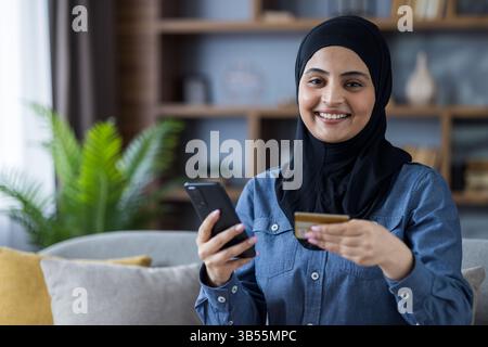 Portrait d'une jeune femme islamique dans un hijab assis sur le canapé à la maison, tenant une carte de crédit et un téléphone portable, regardant la caméra avec le sourire. Banque D'Images