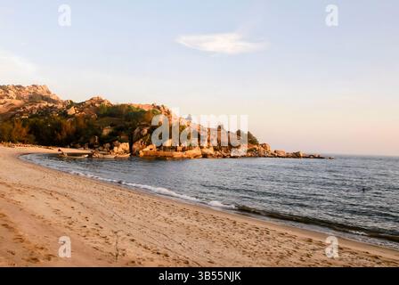Plage tropicale avec vagues calmes, colline rocheuse, bateaux de pêche ancrés et lumière chaude du coucher de soleil sur le rivage en Asie du Sud-est. Banque D'Images