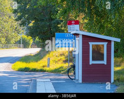 Stockholm, Suède - 6 septembre 2024 : un petit arrêt de bus en bois rouge avec un vélo garé à côté, situé sur une route rurale avec un panneau de direction poi Banque D'Images
