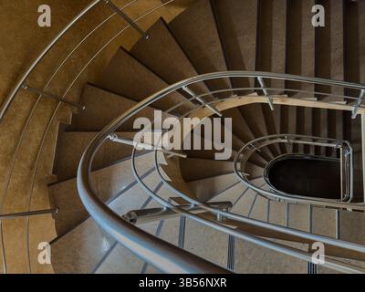 Vue de dessus de l'escalier en colimaçon moderne avec garde-corps en métal et marron à l'intérieur du bâtiment public. Motif géométrique, détail architectural, aucune personne. Banque D'Images
