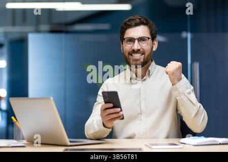 Portrait d'un jeune homme heureux qui est au bureau, assis à un bureau avec un ordinateur portable, tenant un téléphone portable et se réjouissant du succès montre un geste de oui et de victoire, semble satisfait de la caméra. Banque D'Images