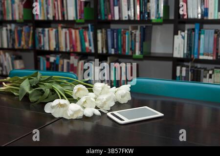 Un bouquet de tulipes blanches et une liseuse sur une table en bois sombre, entouré de chaises sarcelle, avec des étagères remplies de livres en arrière-plan Banque D'Images