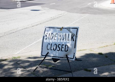 Panneau « trottoir fermé » usé placé sur un trottoir en béton dans une zone urbaine lumineuse, indiquant un détour piéton ou une construction. Banque D'Images