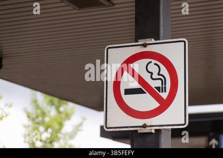 Gros plan d'un panneau d'interdiction de fumer monté sur un poteau sous une structure couverte, indiquant une zone sans fumée dans un espace public. Banque D'Images