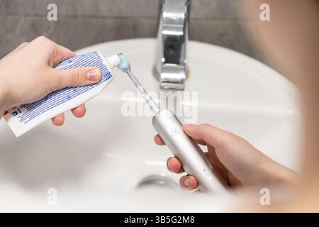 La main d'une personne applique le dentifrice à la tête d'une brosse à dents électrique au-dessus d'un évier, symbolisant l'importance des routines matinales et cohérentes ou Banque D'Images