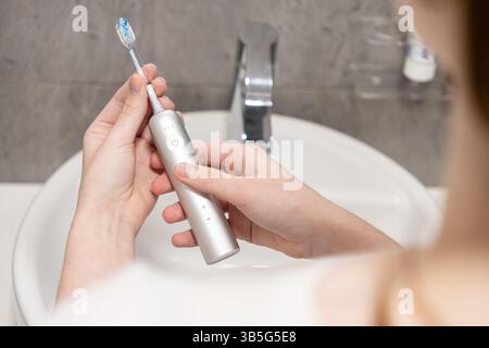 Les mains d'une femme tiennent soigneusement le manche de la brosse à dents électrique et la tête de brosse amovible au-dessus d'un évier, illustrant les étapes de préparation essentielles pour Banque D'Images