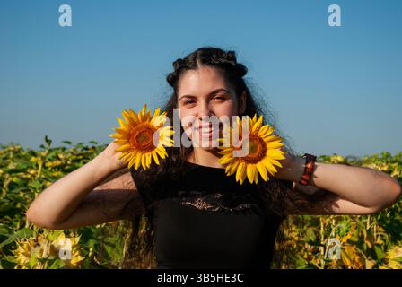 Jeune femme hispanique souriante dans le champ de tournesol tenant des fleurs sous un ciel bleu clair capturant l'essence joyeuse de la nature et de la jeunesse. Banque D'Images