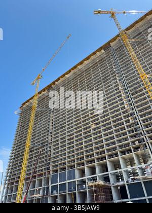 Vue verticale du bâtiment de grande hauteur en construction avec structure en béton et grues à tour contre le ciel dégagé. Développement urbain, pas de personnes. Banque D'Images