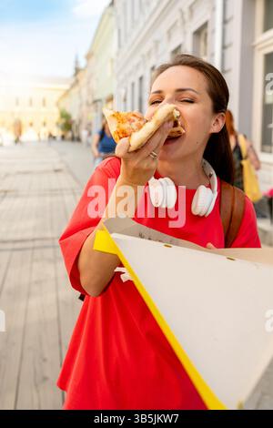 Jeune femme mangeant de la pizza sur le pouce tout en marchant dans la rue en ville Banque D'Images