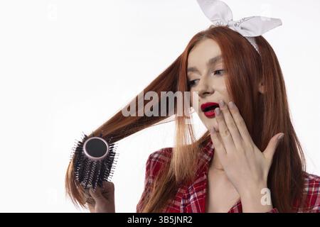 Une jeune femme élégante et à la mode avec des cheveux rouges longs et frappants coiffe habilement ses cheveux avec une brosse Banque D'Images