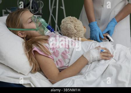 Une fille est allongée dans un lit d'hôpital, recevant des soins du personnel, avec un masque à oxygène et son jouet cher pour le confort Banque D'Images