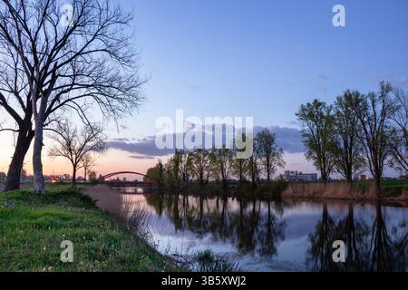 Vue sereine sur la rivière avec une surface d'eau calme montrant des reflets clairs, bordée d'arbres sous un ciel paisible du soir, avec une arche distinctive Banque D'Images