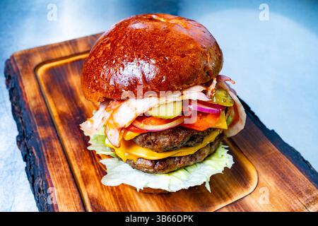 Un appétissant double cheeseburger empilé haut avec du fromage, de la laitue, des tomates, des cornichons, des oignons, et le bacon repose sur une planche de bois. Le pain est doré Banque D'Images