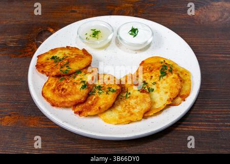 Les crêpes de pommes de terre brunes dorées reposent sur une assiette blanche garnie d'herbes fraîches, accompagnées de deux bols de trempettes crémeuses. Ce plat salé est parfait pour Banque D'Images