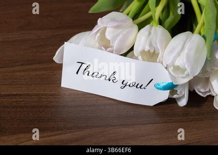 Étiquette cadeau blanche avec message de remerciement et ruban bleu attaché aux tulipes blanches sur une surface en bois foncé, idéal pour exprimer gratitude et appréciation Banque D'Images