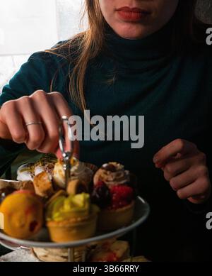 Gros plan des mains d'une femme sélectionnant une petite tarte aux fruits à partir d'un stand à deux niveaux rempli d'un assortiment de desserts miniatures Banque D'Images