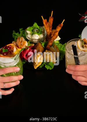 Gros plan de deux mains tenant des cocktails garnis sur une table noire, avec un plateau de hors-d'œuvre mélangés au centre. Banque D'Images