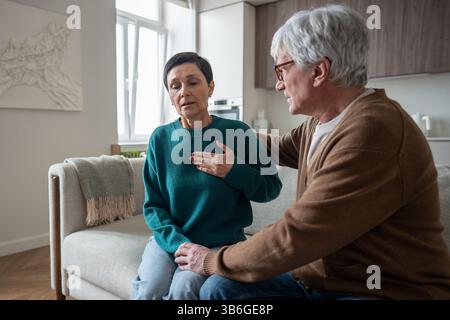 Problèmes de santé de la femme. Femme mature attraper des luttes de poitrine pour l'air réconforté sur le canapé par l'homme de soutien Banque D'Images