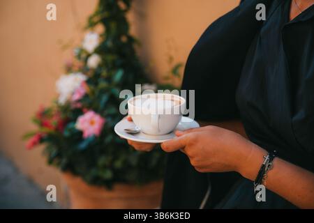 Gros plan de mains tenant une tasse avec un cappuccino en forme de cœur, sur fond de fleurs colorées. La scène transmet une sensation de chaleur et Banque D'Images