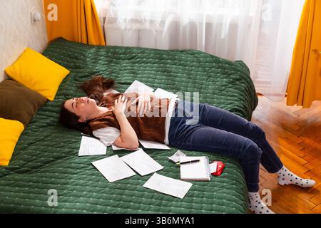 Jeune femme organisant les documents financiers, calculant les coûts mensuels et planifiant le budget du ménage. La femme stressée se trouve sur le lit. Économie et finance Banque D'Images