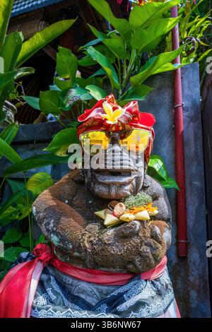 Une statue en pierre balinaise vêtue de sarong traditionnel et de foulard, portant des lunettes de soleil, détient une offrande colorée, entourée de plantes tropicales luxuriantes Banque D'Images