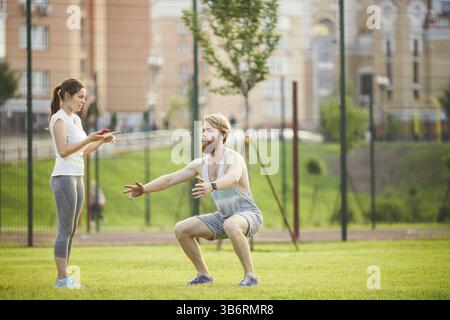Travail d'équipe dans le sport. entraîneur féminin homme de formation avec la barbe dans le parc sur l'herbe verte, pelouse au coucher du soleil. La fille tient le téléphone en main et utilise le chronomètre, g. Banque D'Images