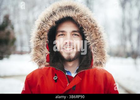 Portrait, close-up of young man smiling avec style avec une barbe habillé en rouge avec une capuche veste d'hiver et fourrure sur sa tête se dresse contre le background Banque D'Images