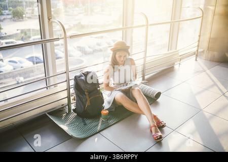 L'attente, retard de transport dans le terminal de l'aéroport ou à la gare. Young caucasian woman in dress and hat siège au tapis avec sac à dos n' Banque D'Images