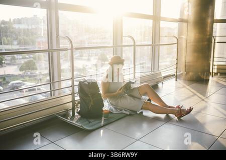 L'attente, retard de transport dans le terminal de l'aéroport ou à la gare. Young caucasian woman in dress and hat siège au tapis avec sac à dos n' Banque D'Images