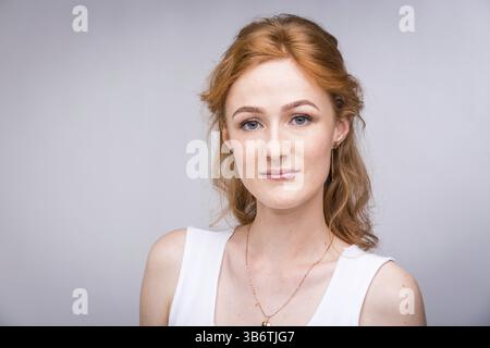 Gros plan portrait jeune, belle femme d'affaires, étudiante avec lred, cheveux bouclés et taches de rousseur sur le visage sur fond gris dans le studio. Habillé de blanc Banque D'Images
