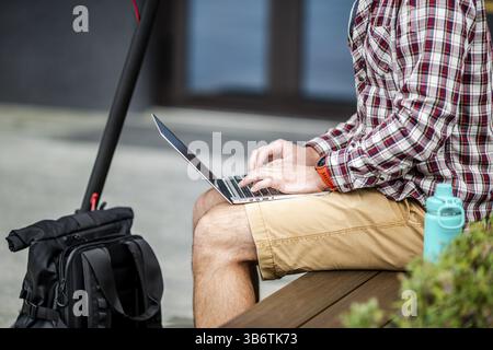 Gros plan de la main de l'homme tapant sur le clavier de l'ordinateur portable tout en étant assis sur le banc en bois dans le centre-ville. Le freelance masculin utilise un ordinateur portable et un scooter électrique. Mes Banque D'Images