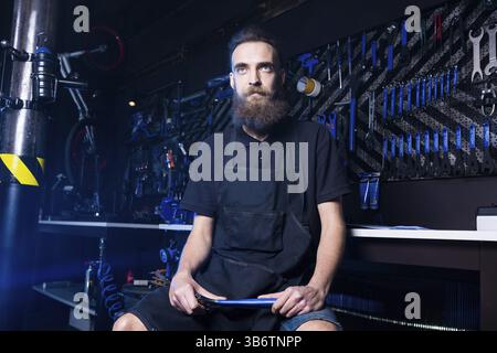 Portrait de propriétaire de petite entreprise de jeune homme à la barbe. Guy atelier mécanicien vélo worker sitting avec l'outil dans sa main dans un vetement noir Banque D'Images