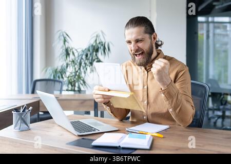 Un homme dans un bureau est excité après avoir lu un document, célébrant un succès ou une bonne nouvelle à son bureau. Banque D'Images