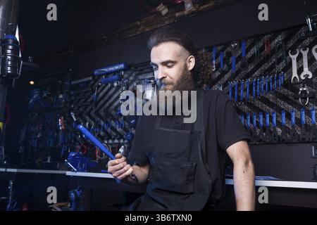 Portrait de propriétaire de petite entreprise de jeune homme à la barbe. Guy atelier mécanicien vélo worker sitting avec l'outil dans sa main dans un vetement noir Banque D'Images