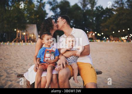 Le concept de vacances en famille. Jeune famille et deux fils assis sur un banc le soir sur une plage de sable. Maman et papa s'embrassent, le frère aîné kis Banque D'Images