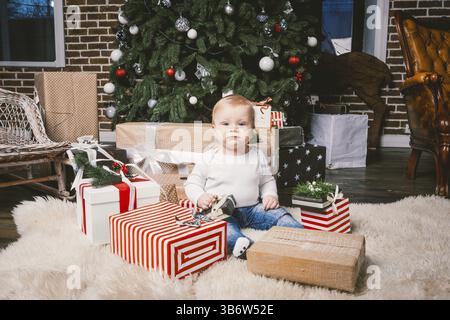 Thème hiver et vacances de Noël. Enfant garçon caucasien blond 1 an assis à la maison près de l'arbre de Noël avec décor du nouvel an sur tapis shaggy Banque D'Images