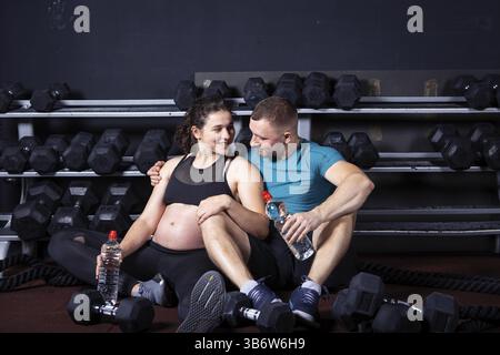 Femme enceinte va faire du sport avec son mari dans la salle de gym pour l'entraînement multidisciplinaire. Couples athlètes attendant bébé tout en se reposant après l'entraînement et les exercices t Banque D'Images