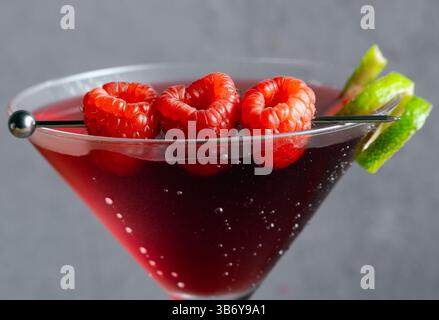 Un gros plan montre un cocktail cosmopolite dans un verre à martini, garni de framboises et d'une touche de citron vert. La boisson est d'une couleur rouge vif. Banque D'Images