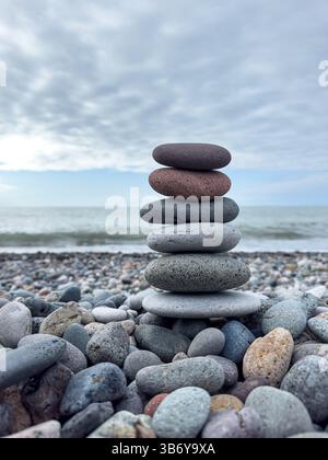 Pile de galets lisses équilibrés sur une plage rocheuse avec la mer et le ciel nuageux en arrière-plan. Composition naturelle, pas de personnes, humeur calme. Banque D'Images