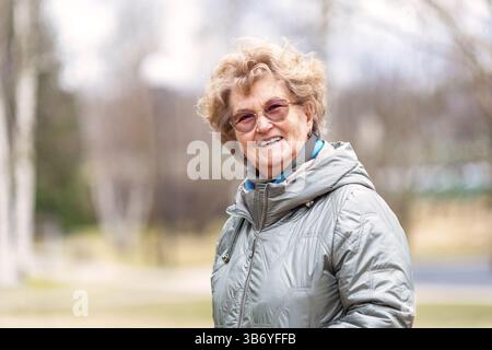 Femme âgée souriante profitant de marcher à l'extérieur au début du printemps, portant une veste chaude et des lunettes, se sentant heureuse, détendue et en bonne santé, montrant une vie active Banque D'Images