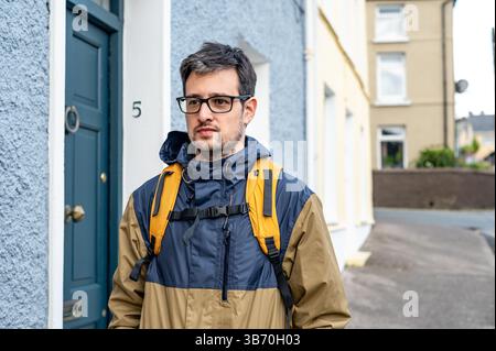 Portrait d'un homme avec des lunettes et un sac à dos jaune debout devant une maison bleue dans une rue irlandaise calme. Banque D'Images