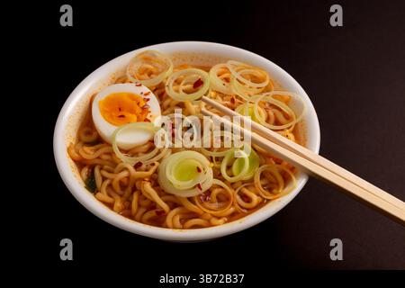 Vue de dessus de la soupe ramen avec nouilles nageant dans un bouillon infusé de piment avec tranches de poireau et œuf, baguettes prêtes à l'action. Fond noir. Banque D'Images