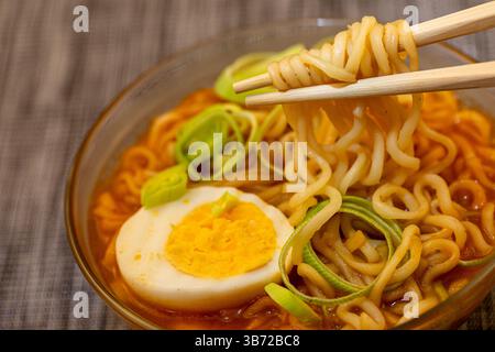 Vue d'angle de ramen épicé avec des fuites vertes et oeufs servis dans un bol en verre avec des baguettes en bois soulevant une partie de nouilles. Banque D'Images