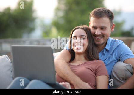 Heureux couple vérifiant l'ordinateur personnel assis dans un canapé dans une terrasse à la maison Banque D'Images