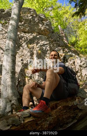 Homme souriant avec un sac à dos assis sur un grand tronc d'arbre parmi les racines exposées sur une pente forestière lors d'une randonnée printanière ensoleillée Banque D'Images