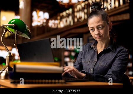 Auteur professionnel et bibliothécaire travaillant sur son ordinateur portable dans une bibliothèque publique, axé sur la fourniture de ressources académiques et de conseils. Évaluer les informations éducatives dans la base de données. Banque D'Images